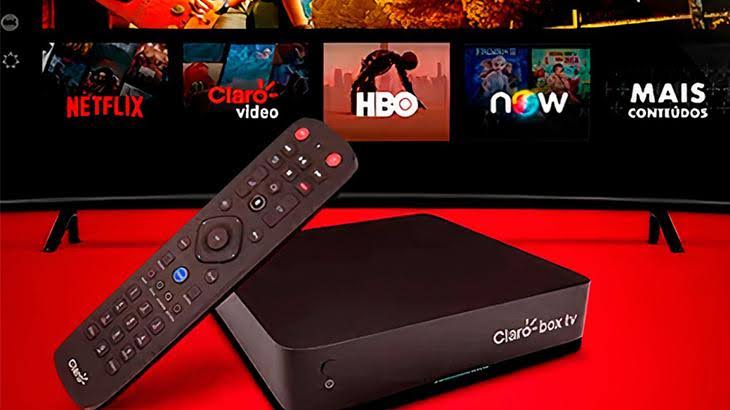 Dispositivo Claro Box TV e controle remoto sobre superfície vermelha, com uma TV ao fundo exibindo aplicativos de streaming como Netflix, Claro Video, HBO e Now.